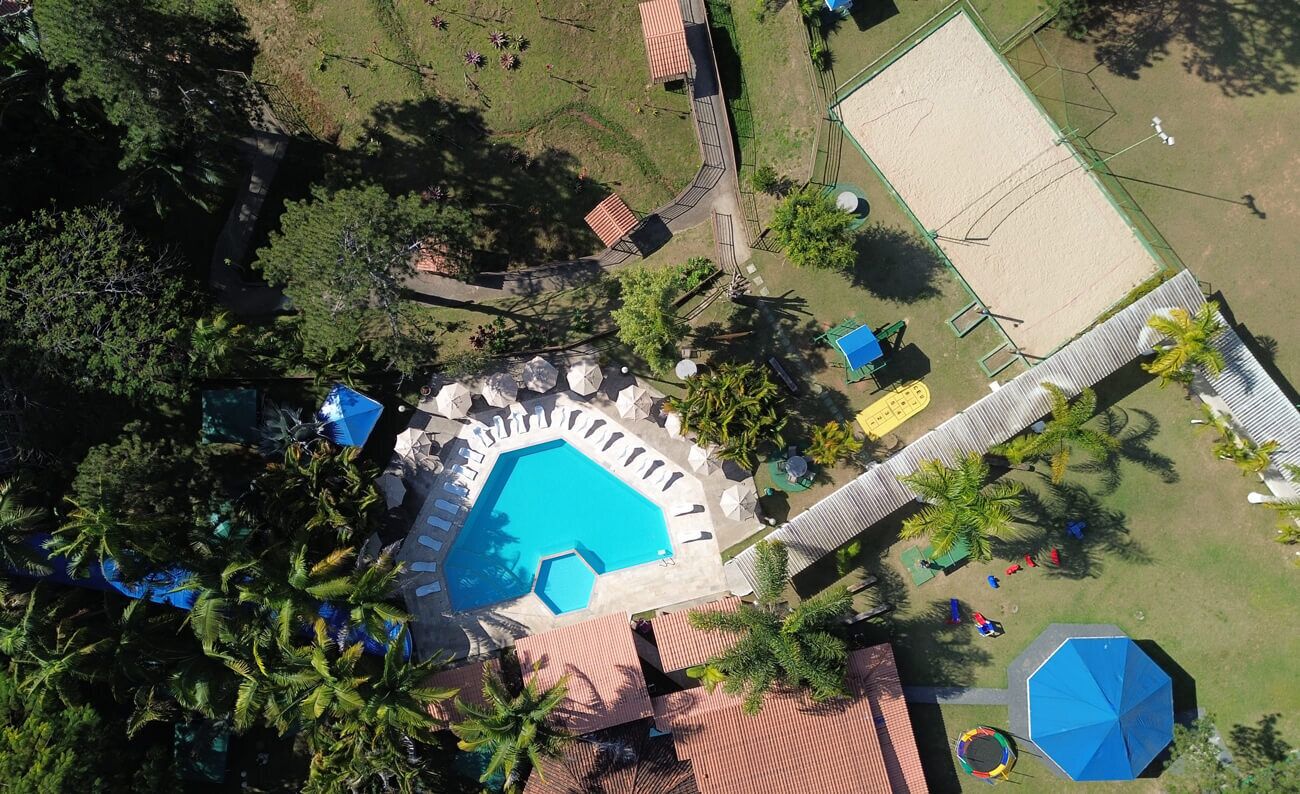 An aerial view of a resort with a central swimming pool, surrounded by palm trees and a tennis court.