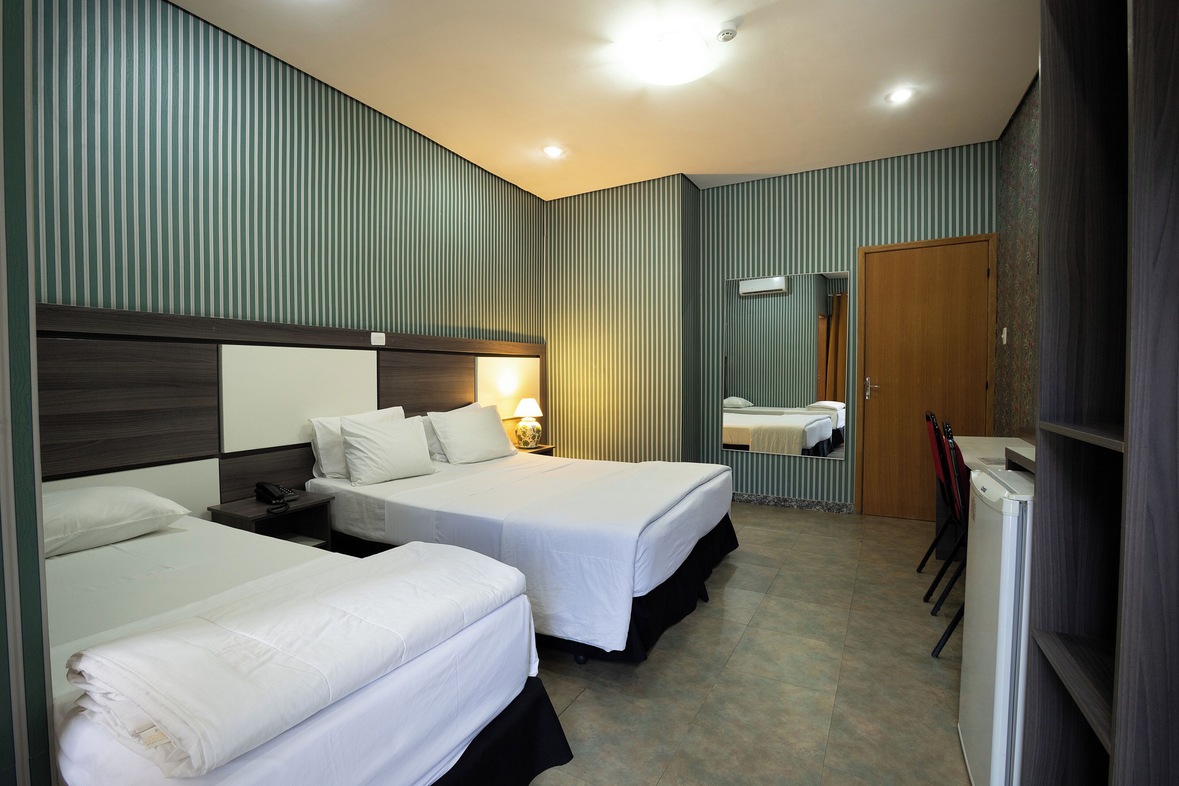 A hotel room with two beds, a nightstand, a desk, a chair, a mirror, and a refrigerator.