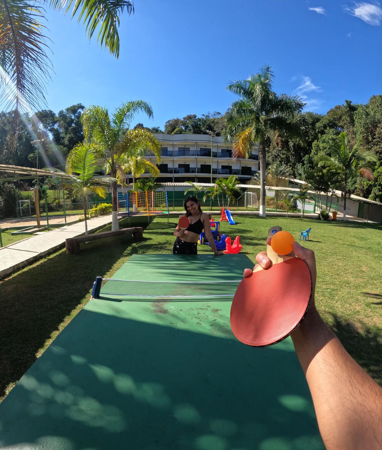 Una cancha de tenis con red, rodeada de palmeras y un edificio al fondo.
