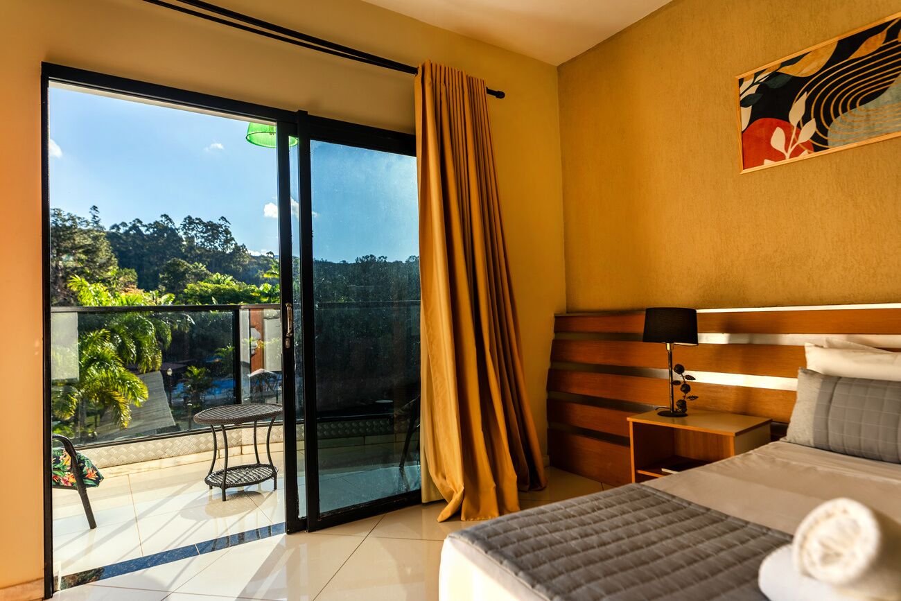 A hotel room with a bed, a nightstand, a lamp, a window with curtains, and a view of greenery outside.