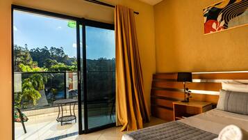 A hotel room with a bed, a nightstand, a lamp, a window with curtains, and a view of greenery outside.