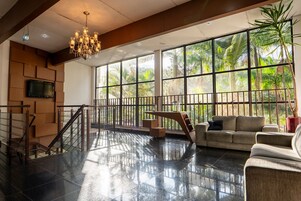 A spacious lobby with a high ceiling, a chandelier, a staircase, a sofa, and large windows offering a view of palm trees.