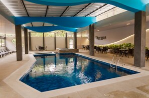 Une piscine intérieure dont les bords sont recouverts de carreaux bleus, entourée de chaises longues et de tables.