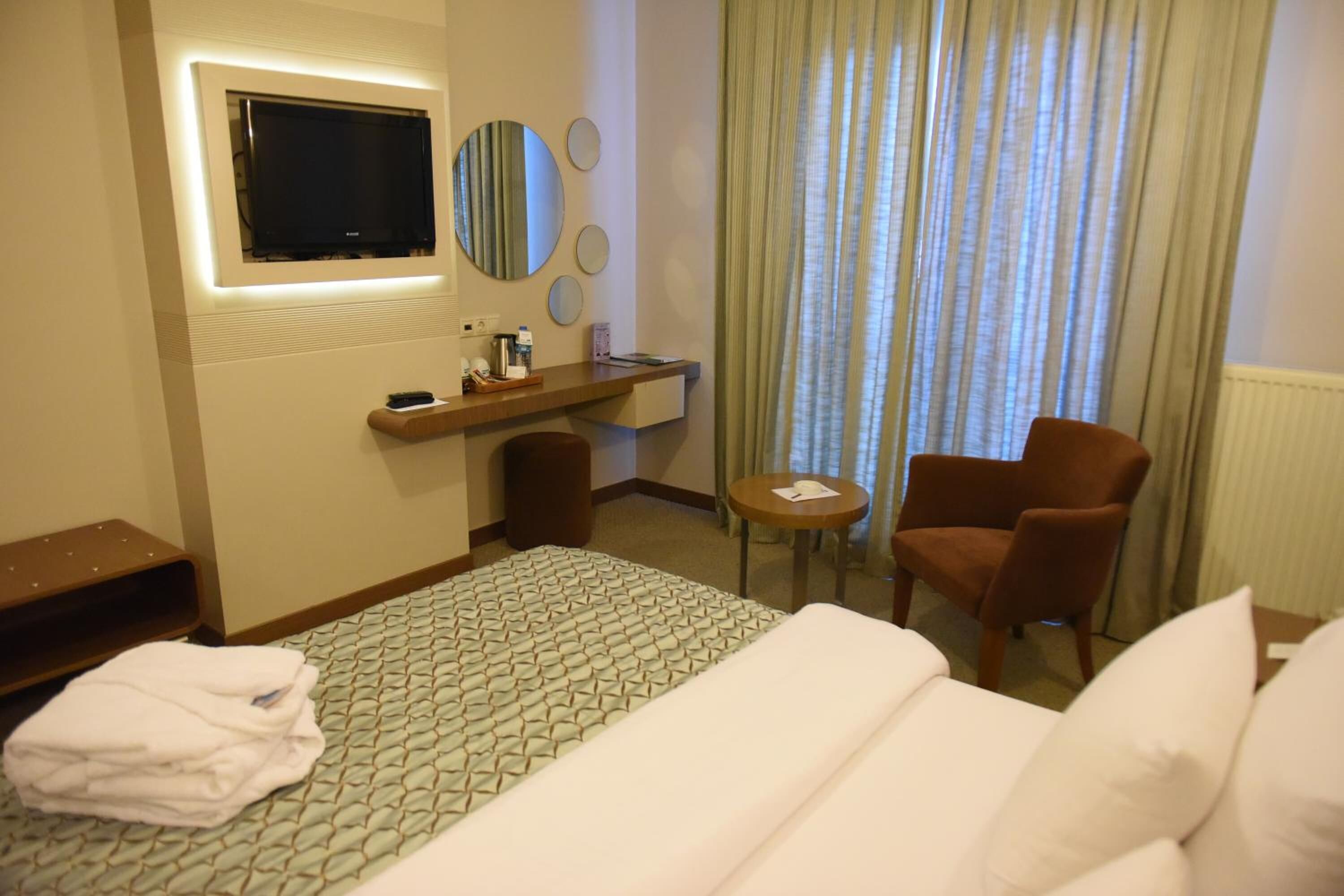 A hotel room with a bed, a desk with a flat-screen TV, a chair, and a small table.