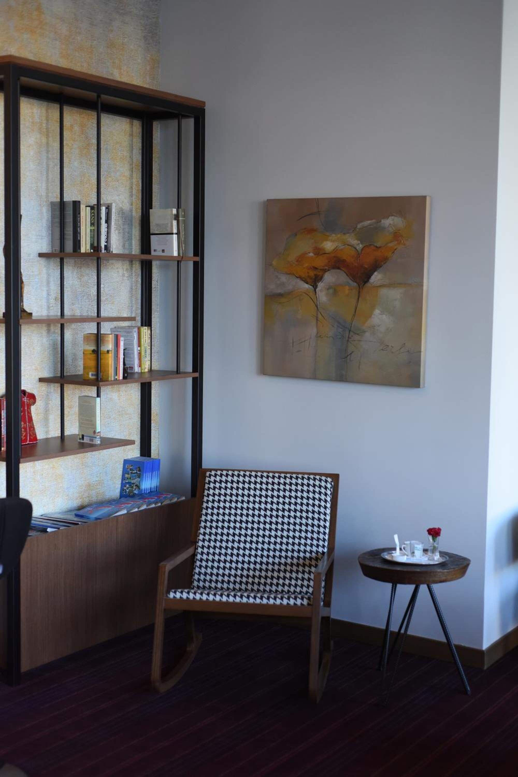 Una habitación con una estantería de madera, un sillón mecedora, una pequeña mesa redonda con un jarrón y un libro, y un cuadro en la pared.
