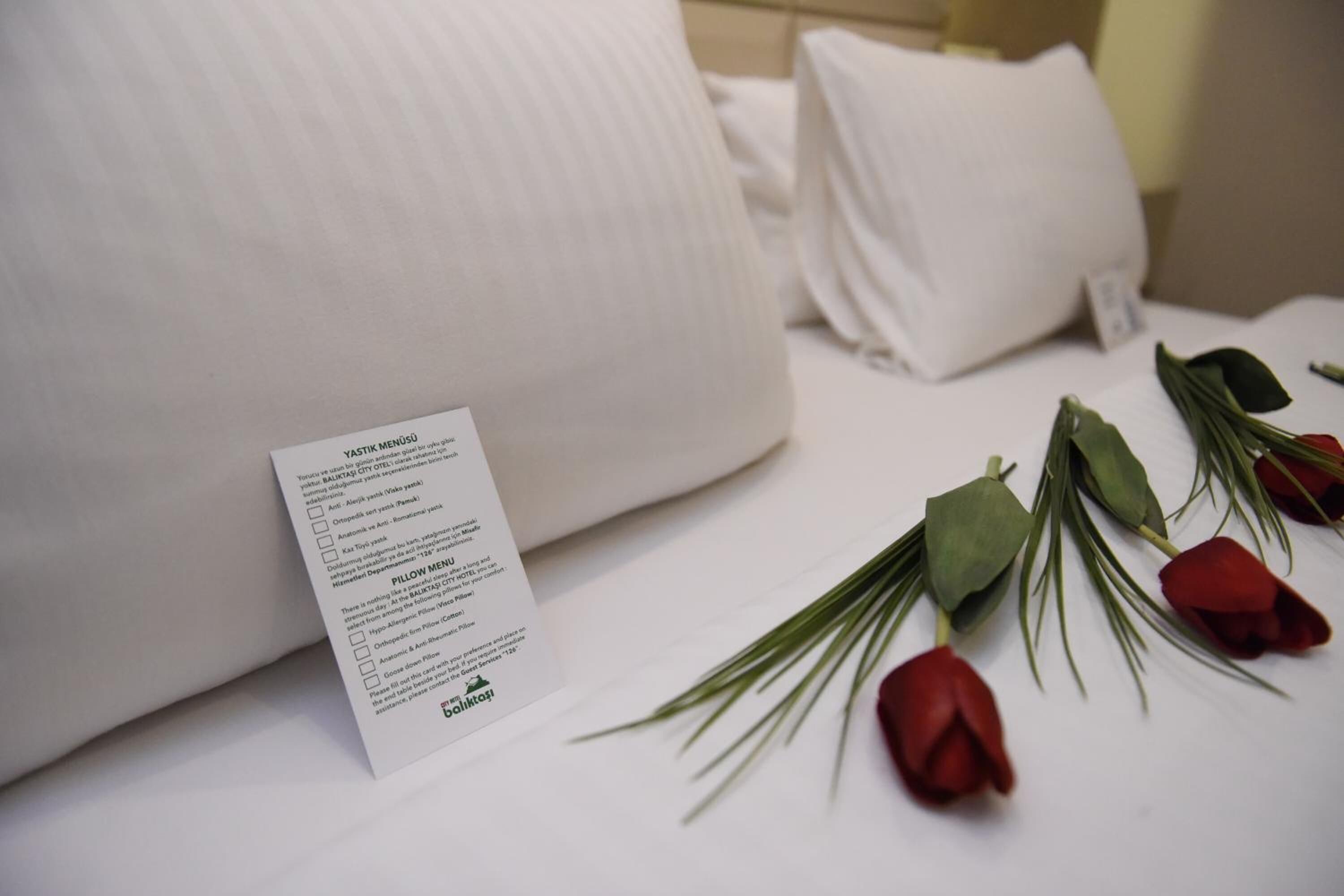 A neatly made bed with white linens, a pillow menu card, and a bouquet of red roses.