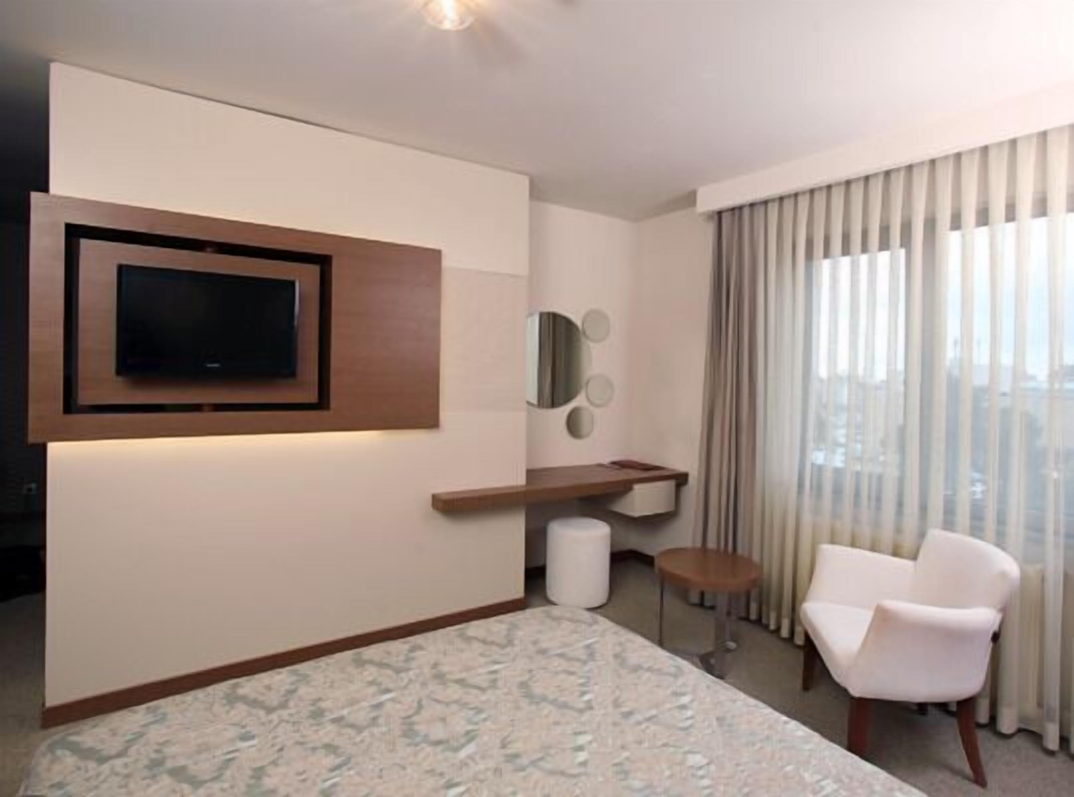 A modern hotel room with a flat-screen TV mounted on the wall, a small round table, a white chair, and a large window with curtains.