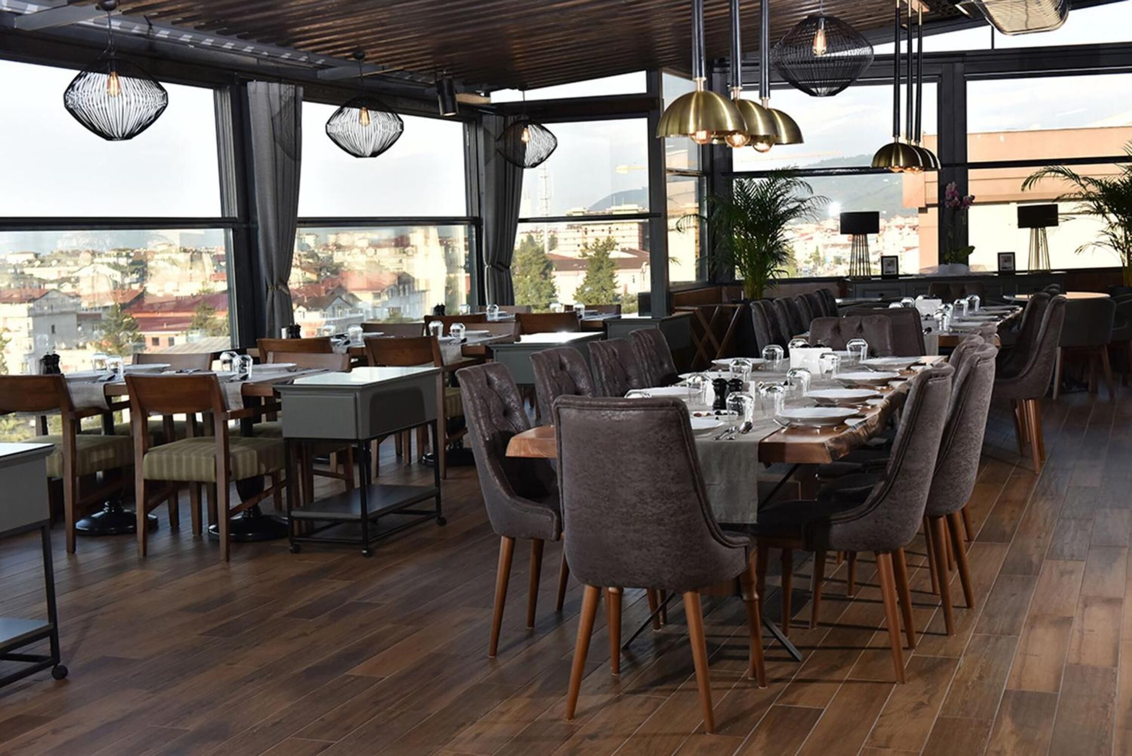 Un restaurante con una larga mesa de comedor preparada para una comida, amplios ventanales con vista a la ciudad y lámparas colgantes.