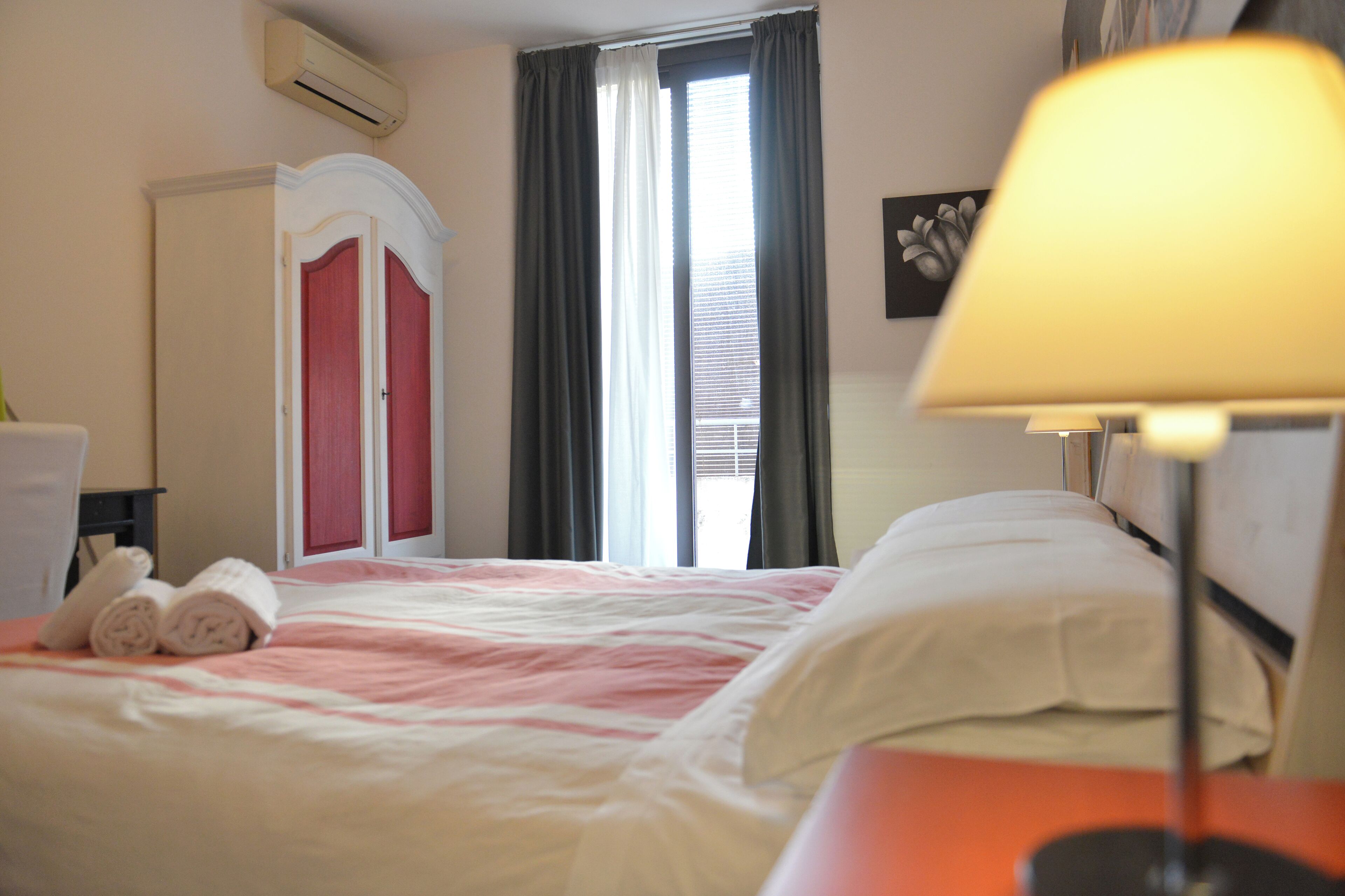 Ein Hotelzimmer mit Bett, Nachttisch, Lampe, einem Fenster mit Vorhängen und einer Tür mit rotem Rahmen.