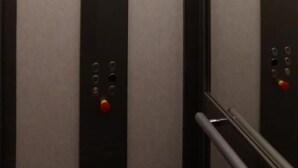 Interior de un ascensor con diseño moderno, revestimiento metálico y panel de control con botones y un botón rojo de emergencia.