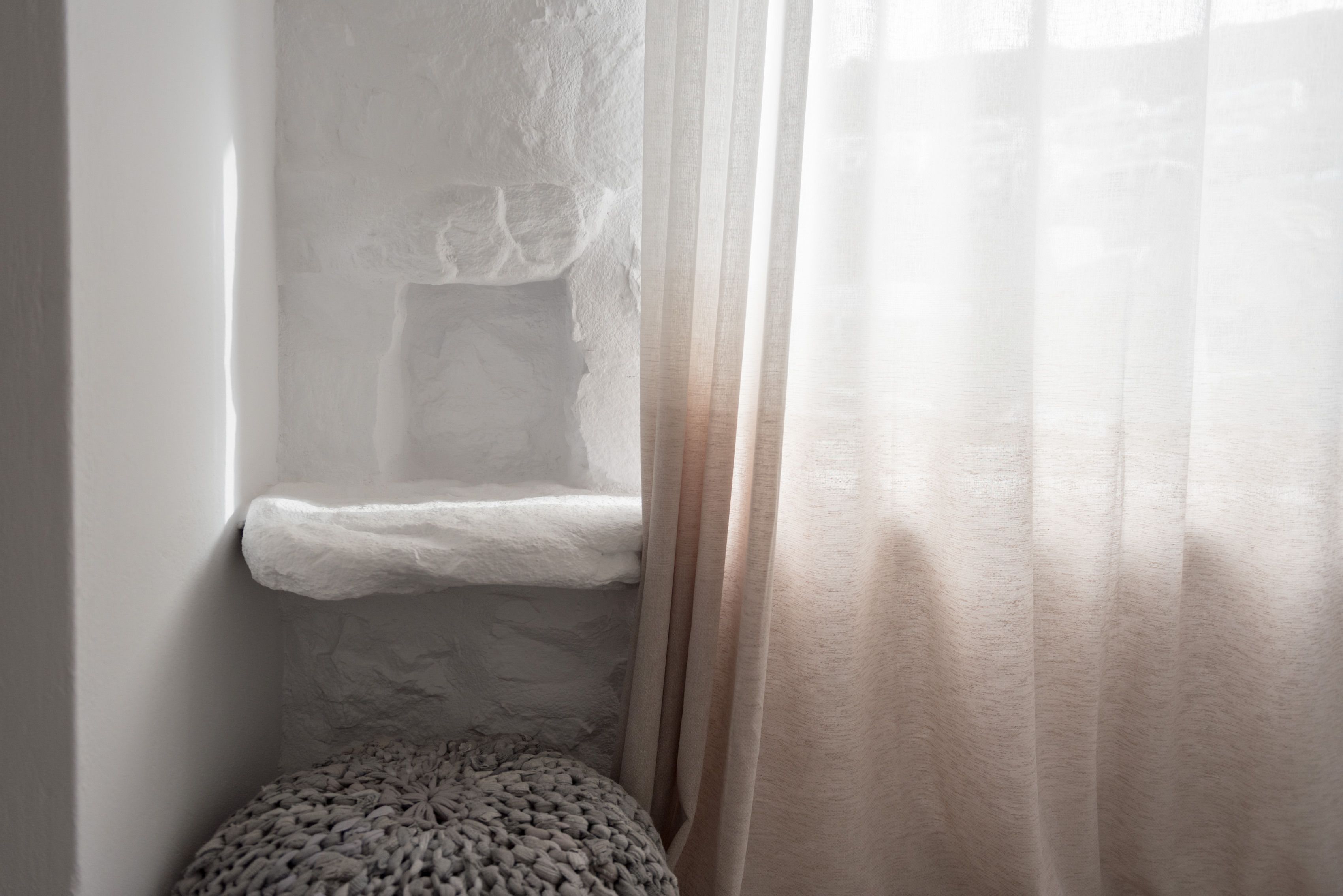 Una esquina de habitación con un banco de piedra, un taburete tejido y cortinas ligeras.