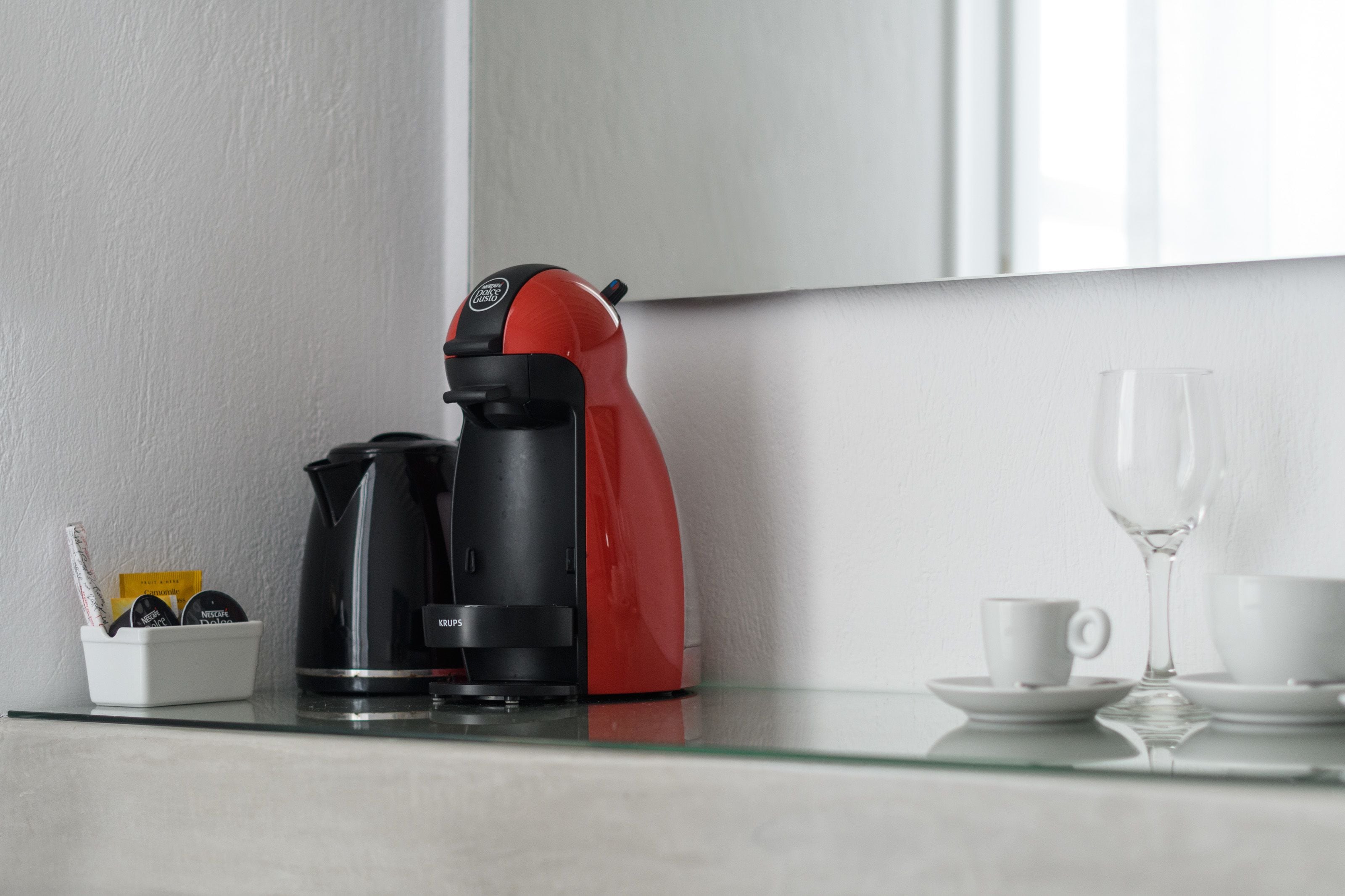 Una cafetera sobre una mesa de vidrio, con una taza y plato, un vaso, y un pequeño recipiente con objetos.