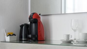 Una cafetera sobre una mesa de vidrio, con una taza y plato, un vaso, y un pequeño recipiente con objetos.