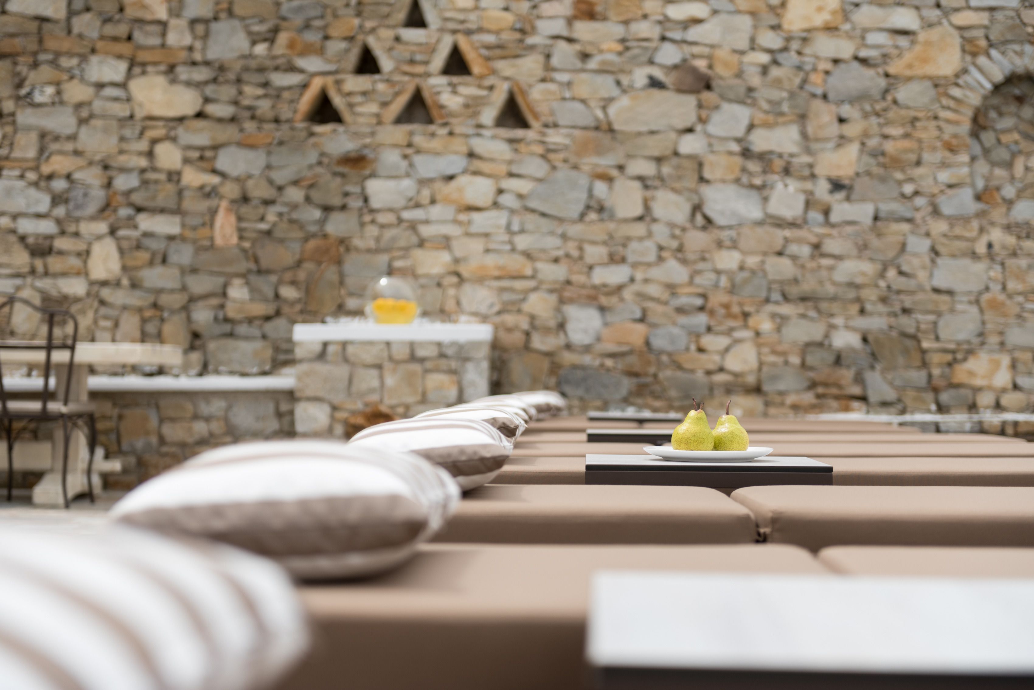 A row of beige cushioned seating with a stone wall backdrop and a small table with a plate of pears.