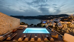 A rooftop infinity pool with lounge chairs, overlooking a coastal town with buildings and a body of water. - Cova Mykonos Suites (Mykonos)