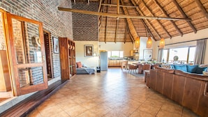 A spacious living area with a high ceiling, a kitchen in the background, and a large window providing natural light. - Dinkweng Safari Camp (Vaalwater)