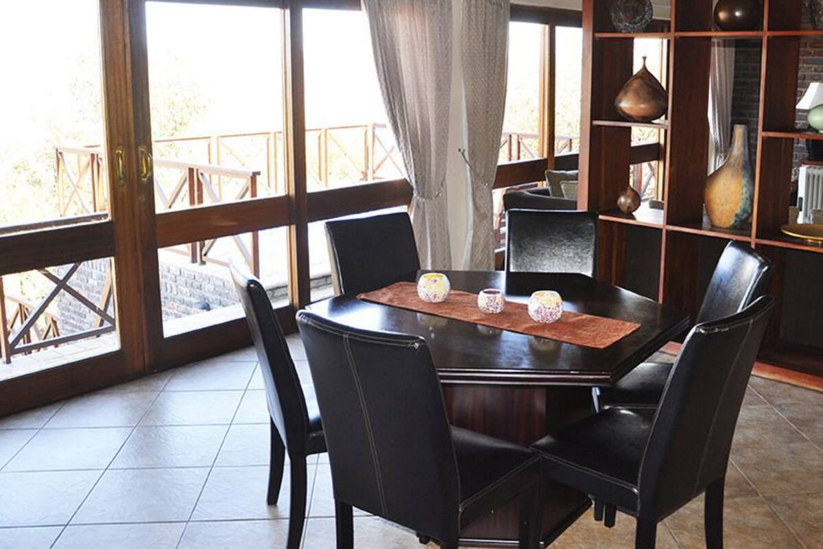 A round dining table with six chairs, a wooden bookshelf with decorative items, and a view of the outdoors through large windows.