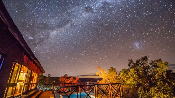 A night sky with stars and the Milky Way over a pool area with a wooden deck and a building with illuminated windows. - Dinkweng Safari Camp (Vaalwater)
