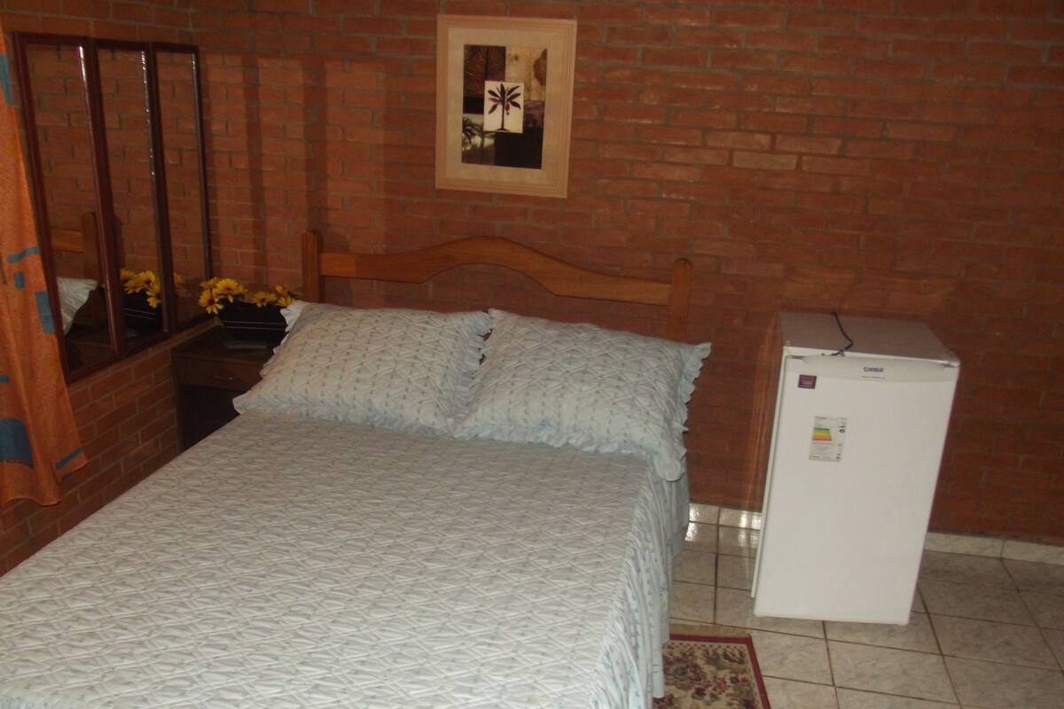 Un dormitorio con cama, cabecera de madera, refrigerador blanco pequeño y pared de ladrillo.