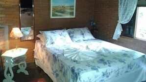 A bedroom with a brick wall, a bed with floral bedding, a bedside table with a lamp, and a painting on the wall. - Pousada Nasa (São Bento do Sapucaí)