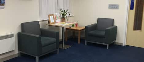 Una habitación con dos sillones verdes, una mesita con una planta, un certificado enmarcado y un cartel que dice 'Rooms 01-12'.