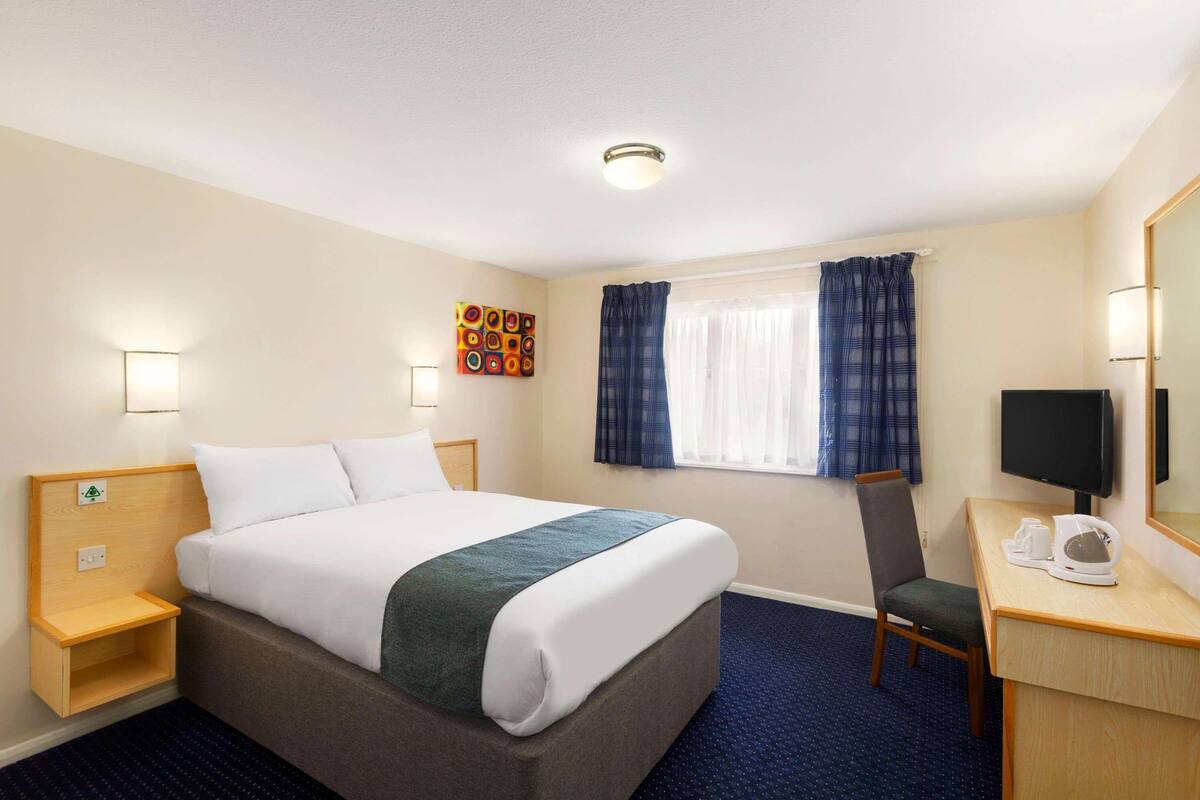 A hotel room with a bed, bedside table, chair, desk, TV, and a window with curtains.