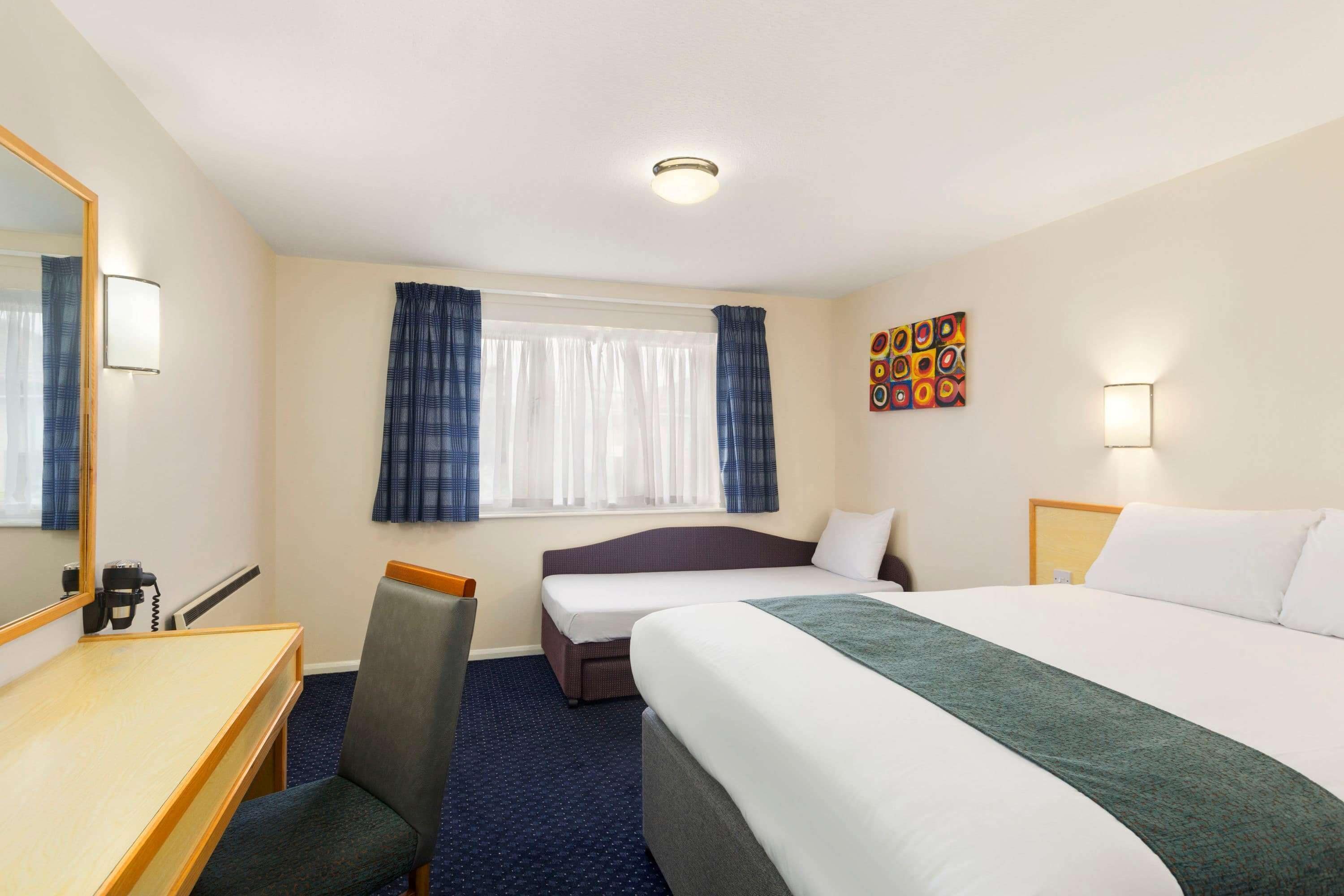 A hotel room with a bed, a desk, a chair, a window with curtains, and a wall with a colorful artwork.