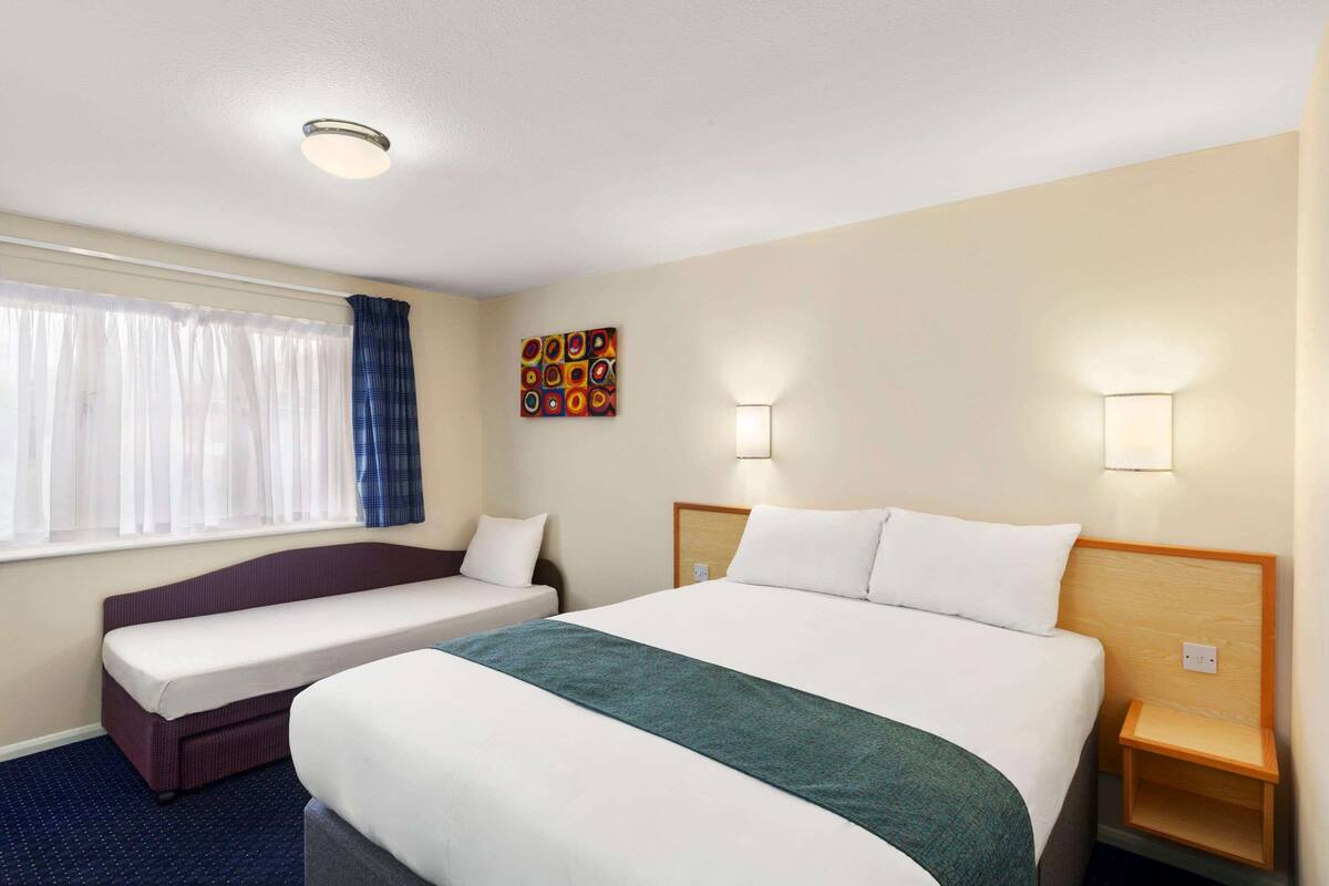 A hotel room with a large bed, a smaller sofa bed, a window with curtains, a wall-mounted artwork, and bedside lamps.