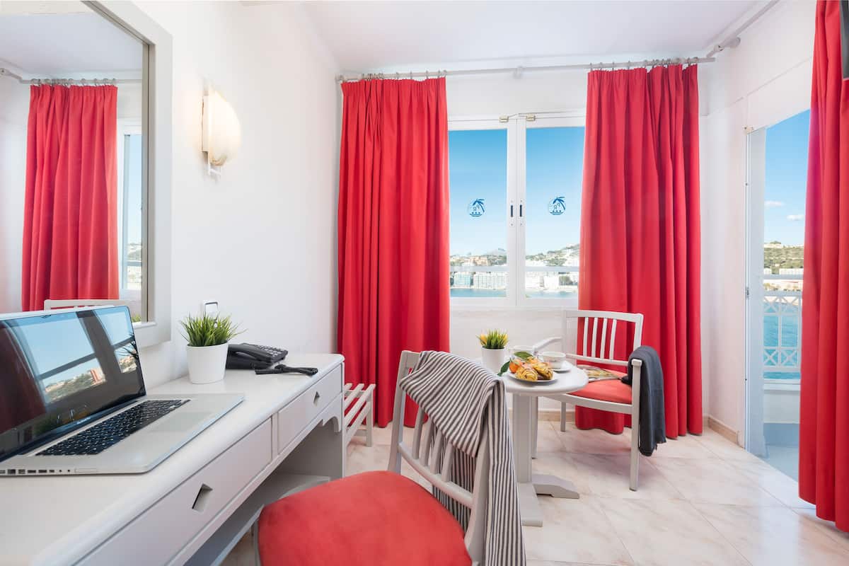 Quarto com um laptop sobre a mesa, uma cadeira, uma pequena mesa com uma planta e uma chávena, e vista para o mar através de grandes janelas com cortinas vermelhas.