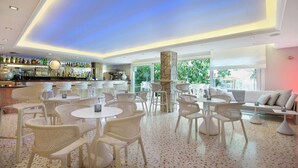 Un espace bar moderne avec des meubles en osier blanc, un comptoir de bar orné de différentes bouteilles et un coin salon comprenant un canapé blanc et des tables rondes.