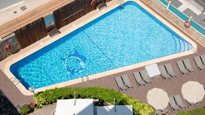 Une piscine rectangulaire, bordée d’une terrasse carrelée, entourée de chaises longues et de parasols, et une clôture en bois en arrière-plan.