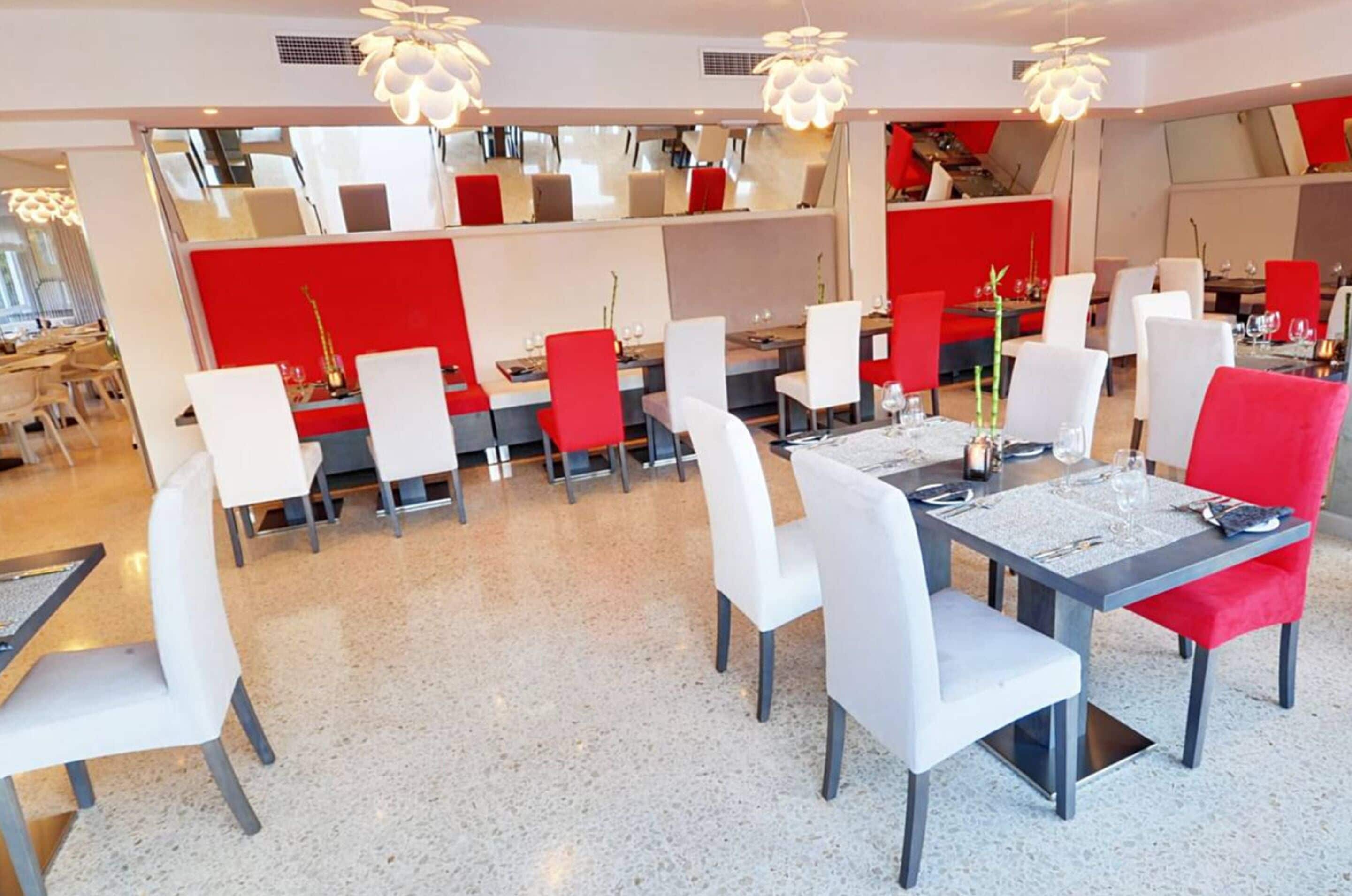 A modern dining area with white and red chairs, tables set with glassware, and pendant lighting.