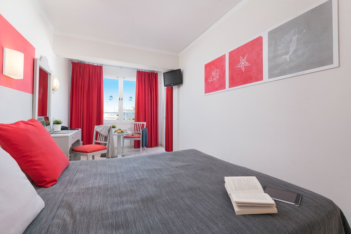 Quarto de hotel moderno com cama cinzenta, almofadas vermelhas, uma mesa com um laptop, uma cadeira e vista da cidade através de grandes janelas.