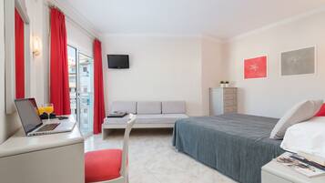 A hotel room with a bed, a desk with a laptop and glasses of orange juice, a red chair, a grey sofa, a television, and two framed pictures on the wall.