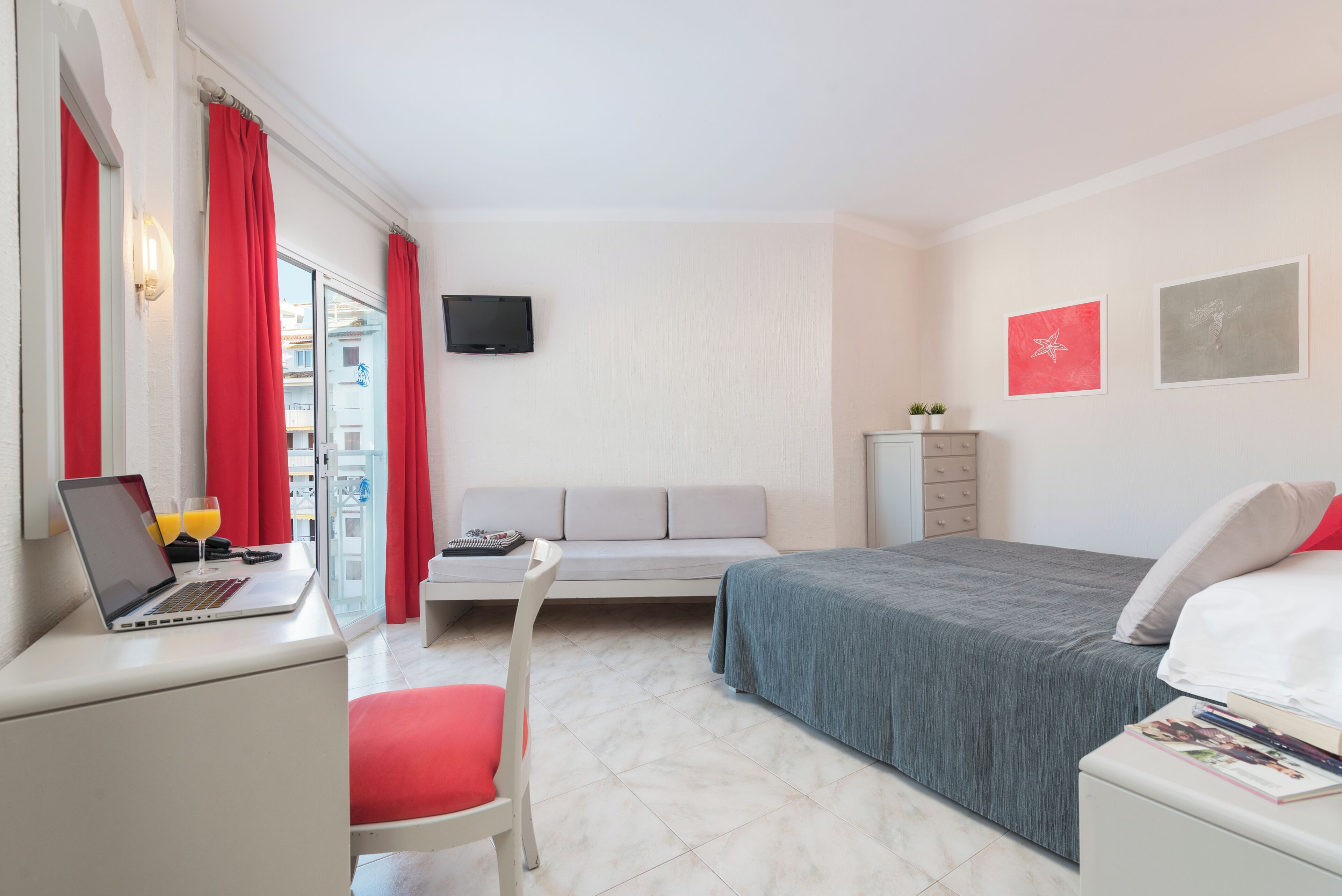 Une chambre d’hôtel avec un lit, un bureau avec un ordinateur portable et des verres de jus d’orange, une chaise rouge, un canapé gris, une télévision et deux tableaux encadrés accrochés au mur.