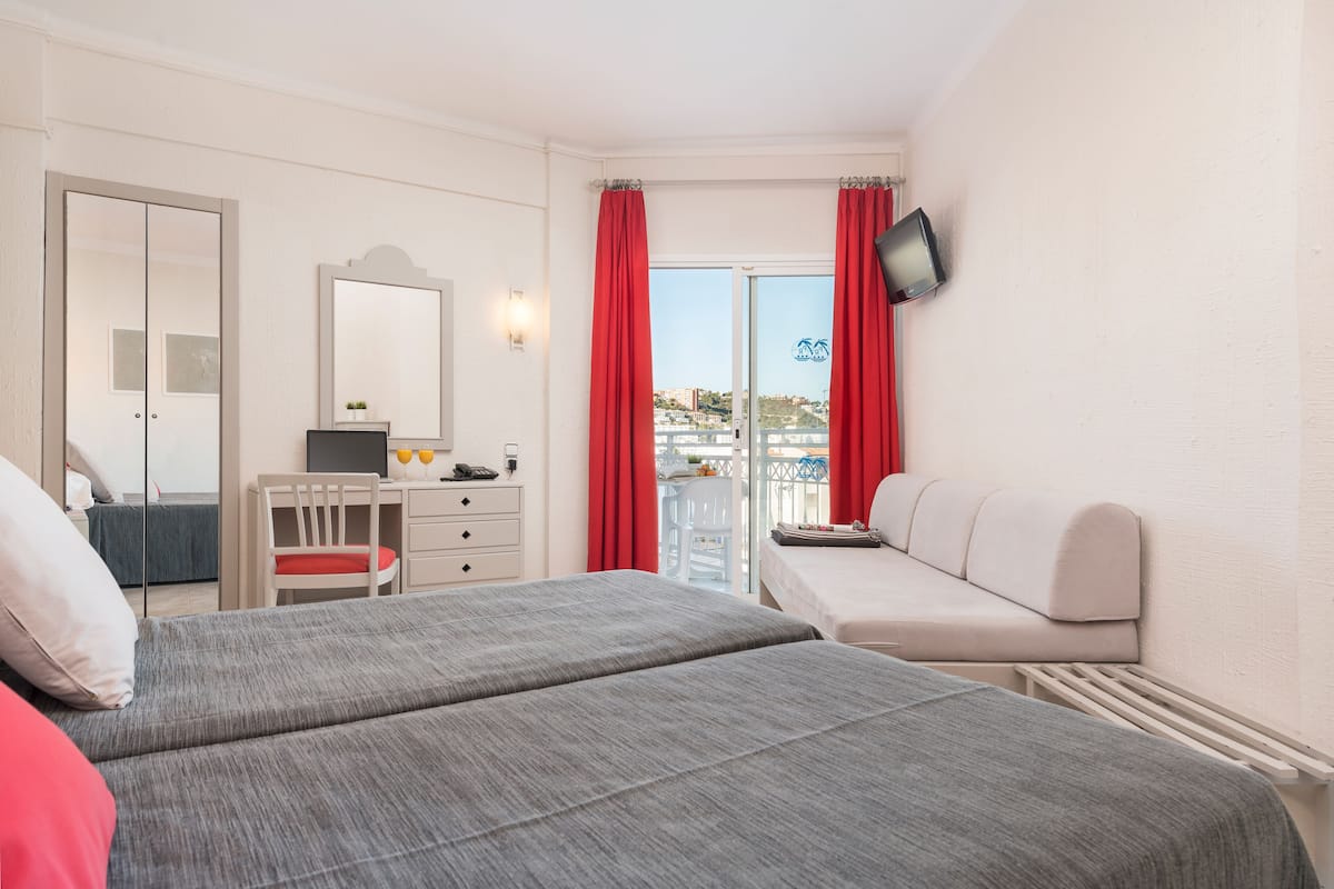 Quarto de hotel com duas camas, uma escrivaninha, uma cadeira, televisão e varanda com cortinas vermelhas.