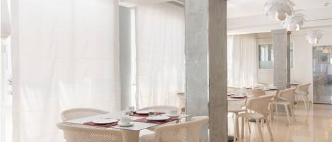 Un vaste espace dédié à la salle à manger, avec des chaises et des tables en osier blanc, de grandes fenêtres ornées de rideaux légers et un éclairage moderne suspendu au plafond.