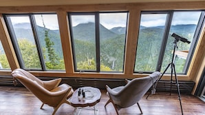 A room with two leather chairs, a small round table, and a telescope on a tripod. Large windows offer a view of a mountainous landscape.