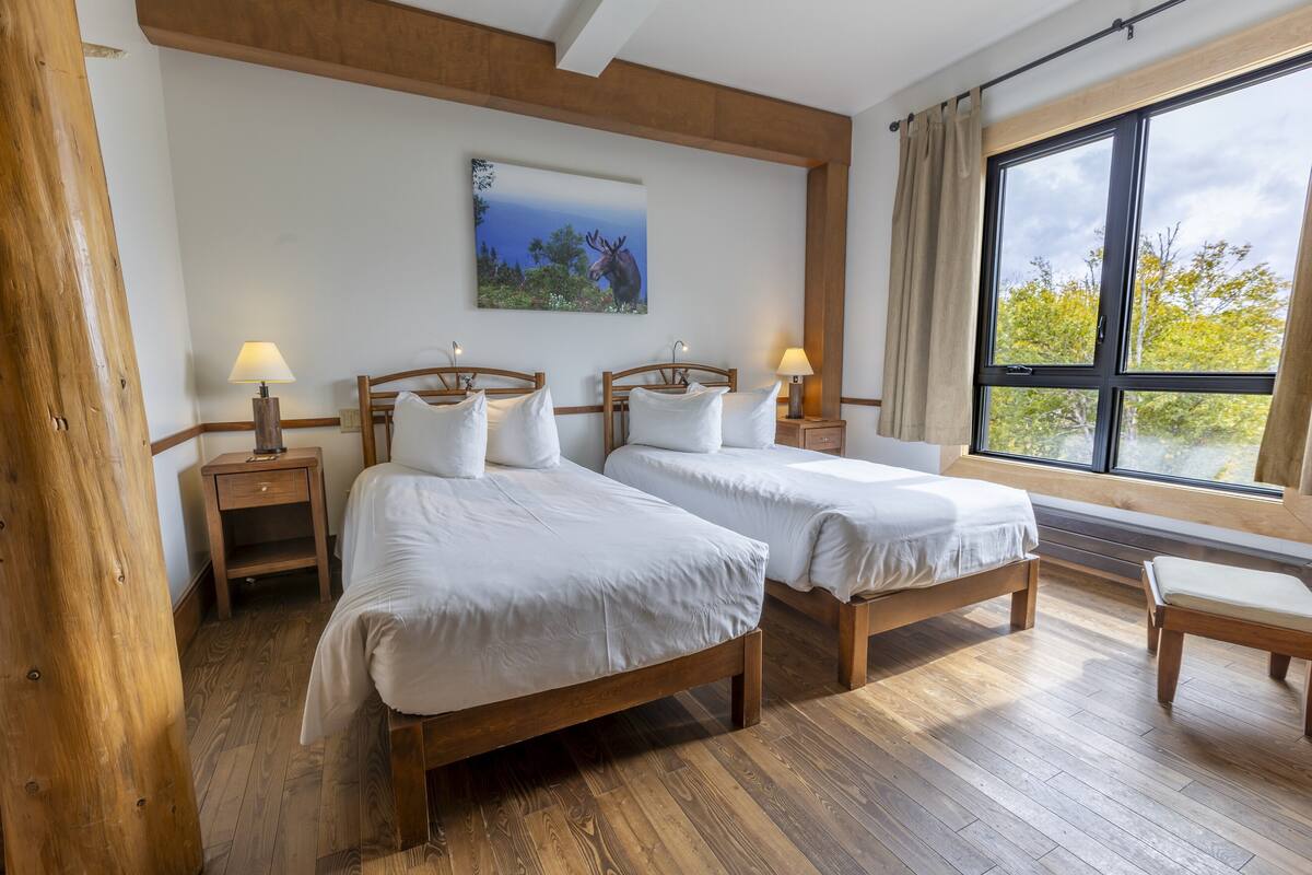 Une chambre d’hôtel avec deux lits en bois, une grande fenêtre donnant sur des arbres et un tableau représentant un élan accroché au mur.