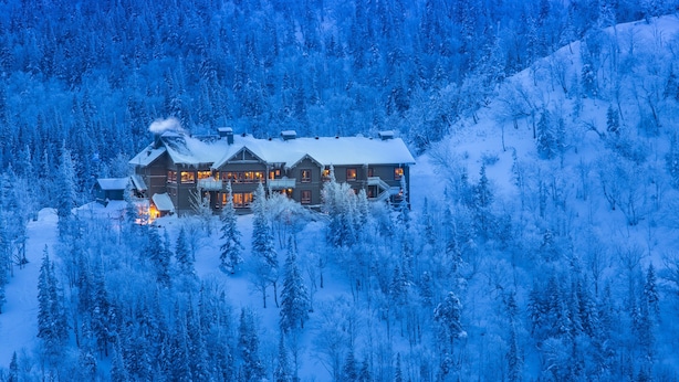 A large, multi-story lodge with a snow-covered roof, surrounded by dense forest.