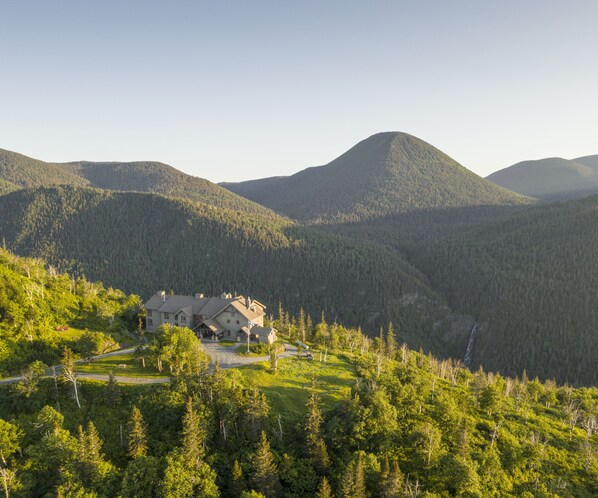 A large house with a spacious driveway and a well-maintained lawn, surrounded by dense forest and hills. - Auberge de Montagne des Chic-Chocs - Sepaq (Sainte-Anne-des-Monts)