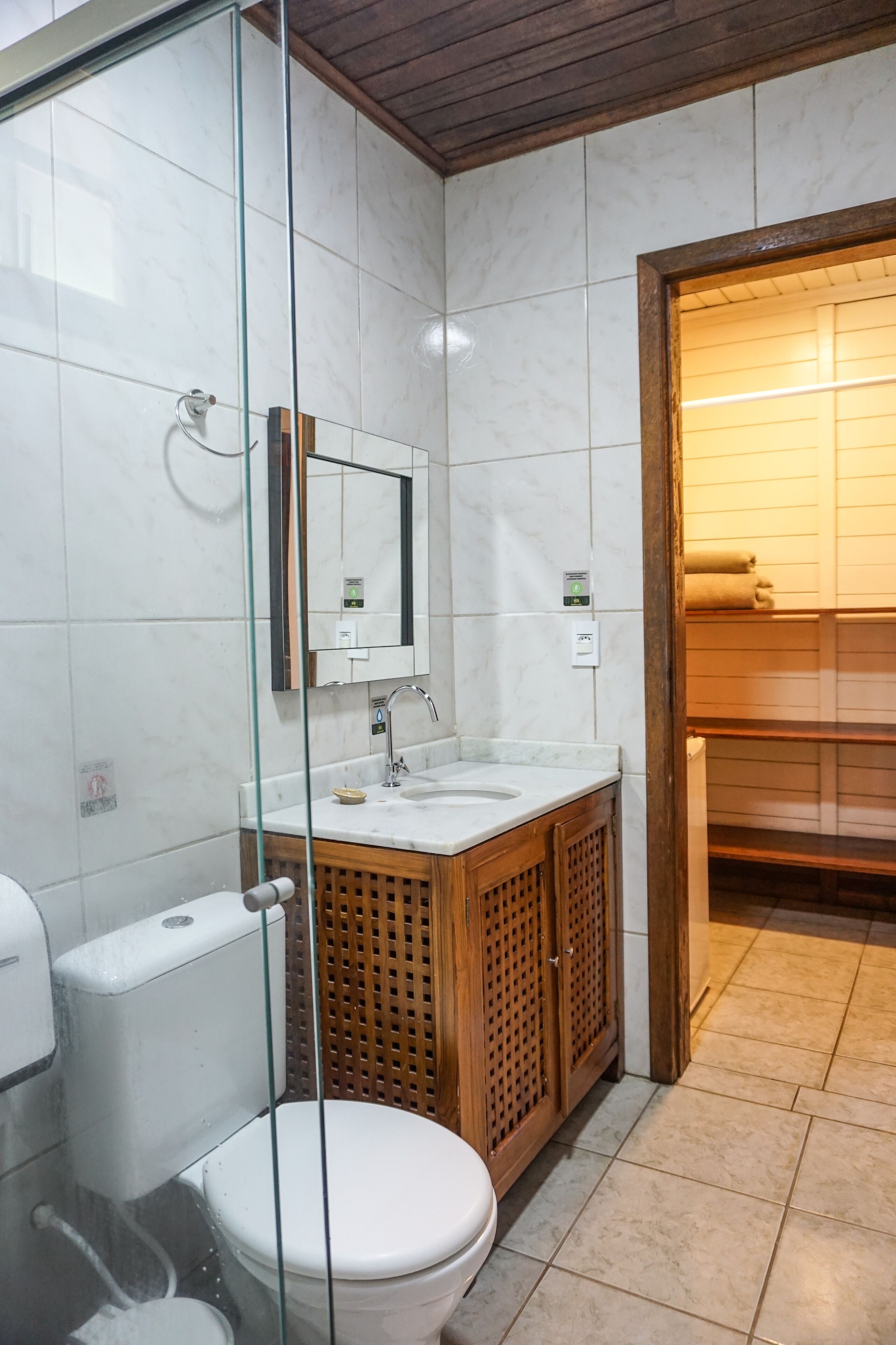 A modern bathroom with a glass shower enclosure, a white toilet, a wooden vanity with a mirror, and a tiled floor.