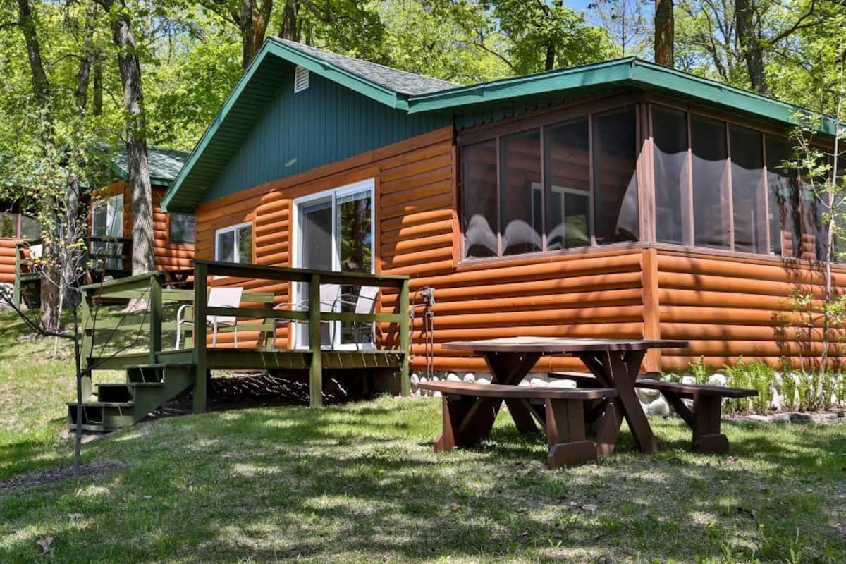 Una cabaña rústica de troncos con techo verde, una terraza de madera y una mesa de picnic.