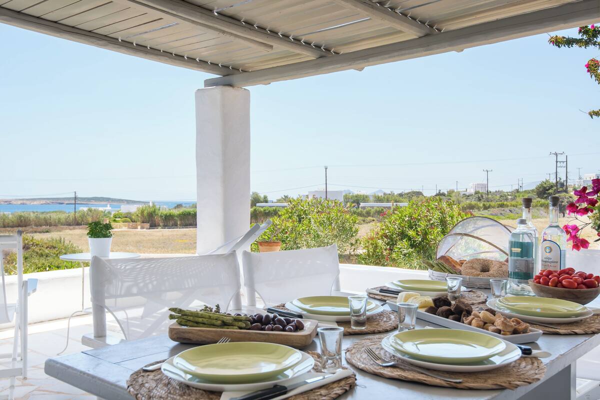 Un área de comedor al aire libre con vistas al mar, muebles blancos y una mesa dispuesta con platos, vasos y comida.