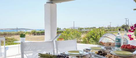 An outdoor dining area with a view of the sea, white furniture, and a table set with plates, glasses, and food.