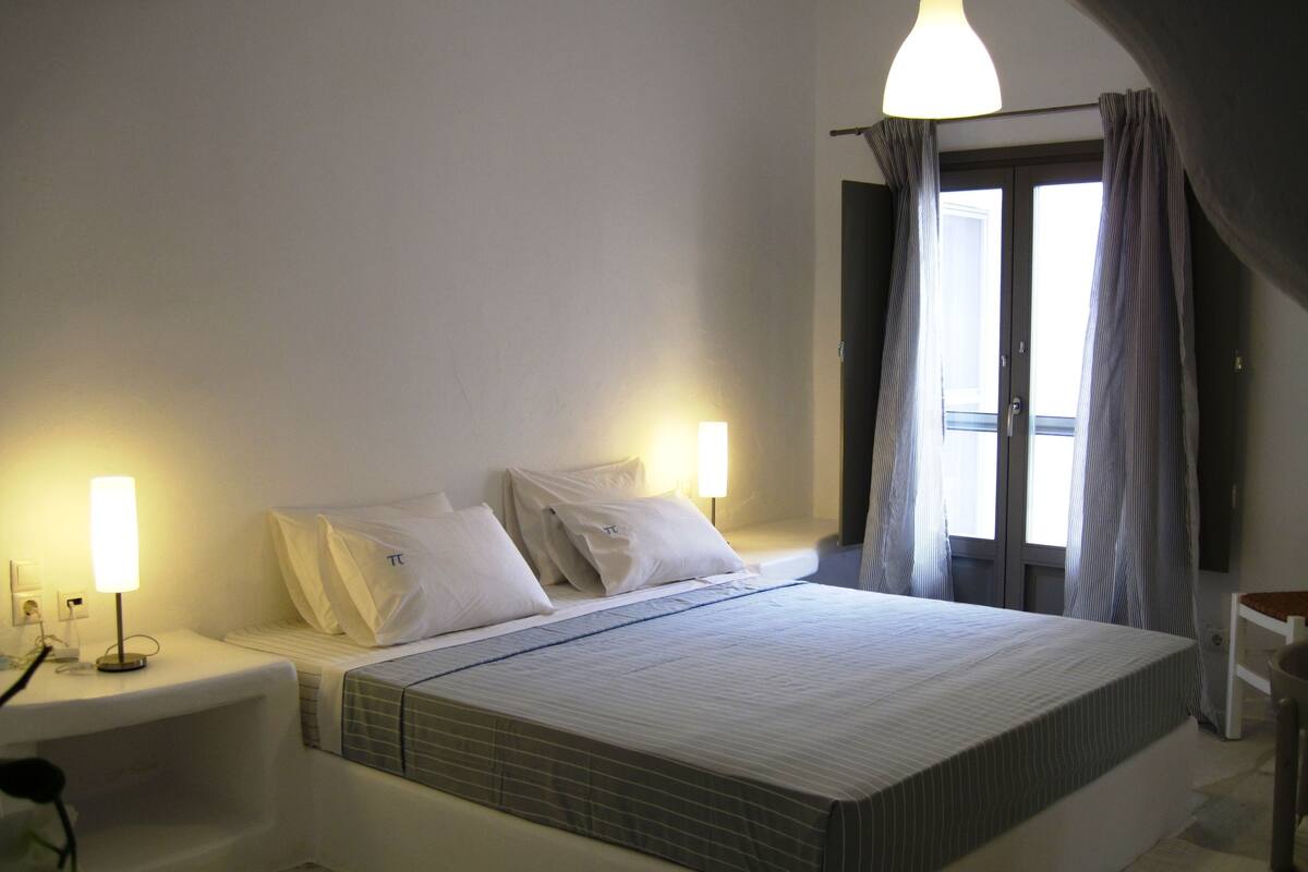 Un dormitorio con una cama grande, dos lámparas de noche, una ventana con cortinas y una pequeña mesa.