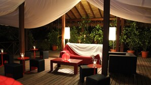 Een terras met houten tafels en stoelen, een overdekt gedeelte met een rode bank, en potplanten.