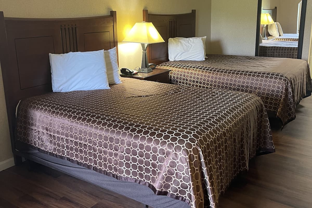 Une chambre d’hôtel avec deux lits, chacun agrémenté d’une couvre-lit à motifs et d’une tête de lit en bois. On y trouve une table de chevet avec une lampe et un téléphone. La chambre est dotée d’un plancher en bois et d’un miroir au mur.