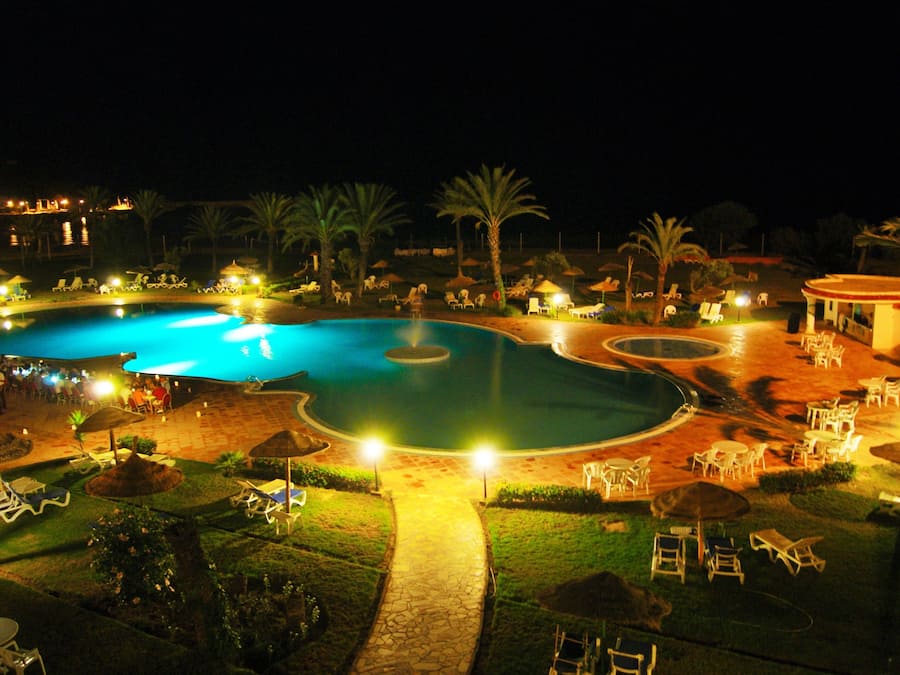 Nattebillede af et resortområde med flere pools, liggestole og parasoller.