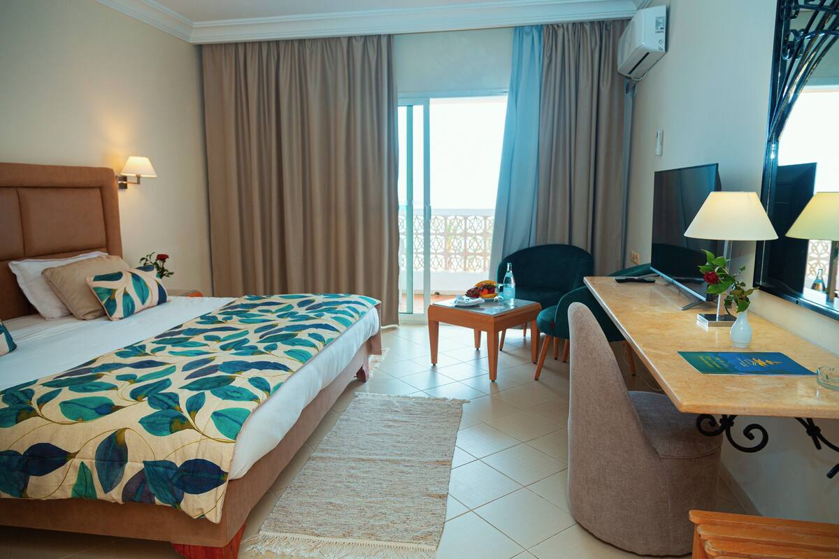 A hotel room with a bed, a desk with a chair, a TV, and a window with curtains.
